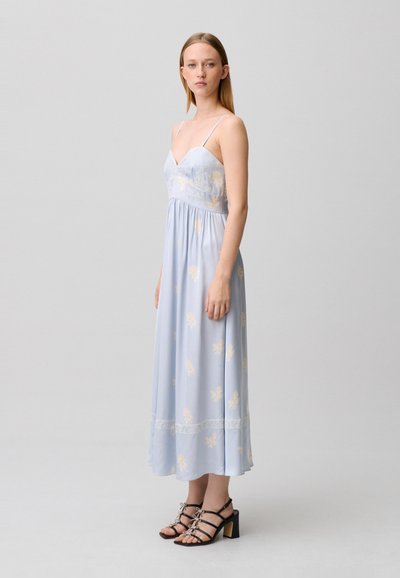 Giovane donna che indossa un maxi abito a fiori azzurro chiaro con spalline sottili e sandali neri con tacco, in piedi davanti a uno sfondo grigio uniforme.