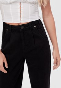 Black high-waisted pants featuring front pleats, a button closure, and side pockets, made from a sturdy cotton blend fabric.