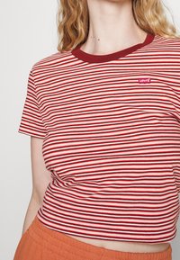 Kobieta w czerwono-białą w paski koszulce Levi's z czerwoną kołnierzykiem, w połączeniu z pomarańczowymi spodniami o wysokim stanie, ściągniętymi na wysokości połowy torsu.