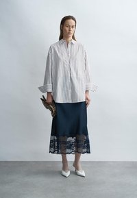 Gestreifte Bluse mit langen Ärmeln in Weiß und Marineblau, kombiniert mit einem marineblauen Rock mit Spitzenbesatz. Weiße spitze Schuhe und eine Clutch mit Leopardenmuster vervollständigen den Look.