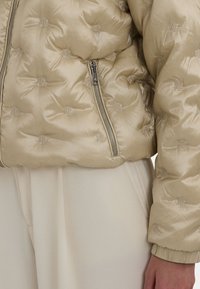 Beżowa kurtka puchowa z błyszczącego materiału, z wzorem tonalnego logo, zamkiem błyskawicznym i ściągaczami, podkreślająca pikowany design.