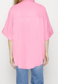 Rosa oversized skjorta med korta ärmar och rundad nederkant, tillverkad av lätt material. Baksidan har en yoke-detail.