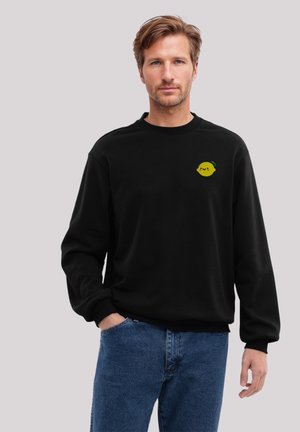 Schwarzer Sweatshirt mit langen Ärmeln, gerippten Bündchen und einem lächelnden Zitronen-Graphic auf der linken Brust. Kombiniert mit blauen Jeans.