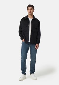 Zwarte button-up shirtjas gemaakt van stof met twee borstzakken, gecombineerd met blauwe jeans en witte sneakers, met een relaxte pasvorm.