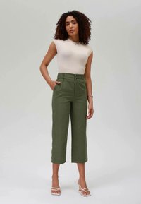 Zaļas apgrieztas bikses ar piegriezumu, augstu jostu un priekšējām kabatām. Apvienotas ar gaiši bēšas ribotu bez piedurkņu topu un baltām sandālēm.