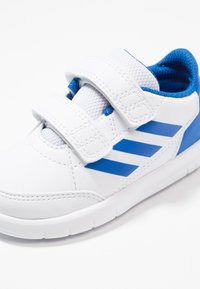 Vit sneaker med blå detaljer, som har en slät syntetisk ovandel, nät-tunga och kardborreband för stängning. Gummisula med mönster på sulan.