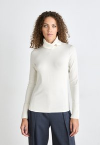 Cremefarbener Rollkragenpullover mit gerippten Bündchen, der ein Broschendetail am Hals aufweist. Weicher Stoff mit einer taillierten Silhouette. Kombiniert mit dunklen Hosen.