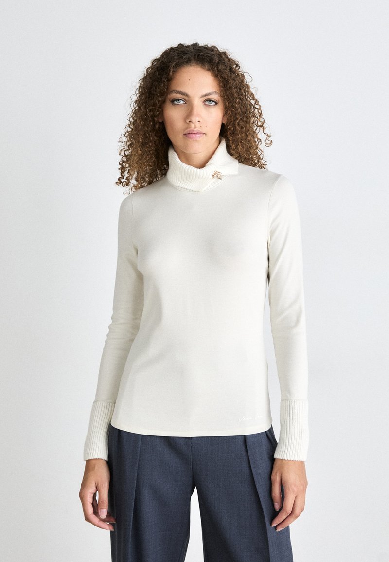 Cremefarbener Rollkragenpullover mit gerippten Bündchen, der ein Broschendetail am Hals aufweist. Weicher Stoff mit einer taillierten Silhouette. Kombiniert mit dunklen Hosen.