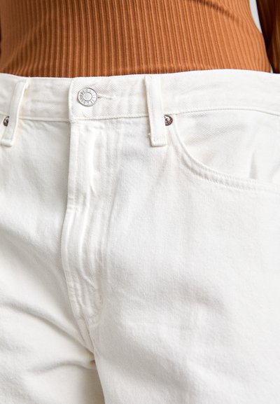 Jeans en denim blanc avec une finition texturée, dotés d'un bouton argenté et de coutures contrastées. La taille présente des passants de ceinture et des poches.