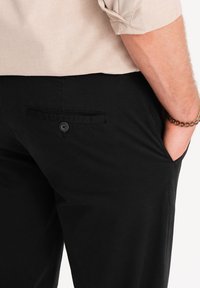 Pantalones negros con bolsillo trasero y detalle de botón, hechos de tela lisa. Una camisa crema es parcialmente visible en la cintura.