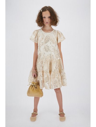 Κορίτσι που φοράει εκρού δαντελένιο φόρεμα και χρυσά παπούτσια, κρατώντας μια χρυσή τσάντα με κορδόνια, σε απλό λευκό φόντο.