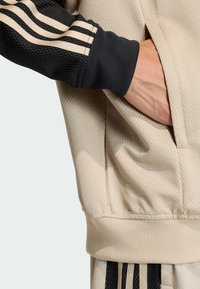 Beež athletikajakk tekstureeritud kangaga, millel on mustad võrgusarnased varrukad ja kolm musta triipu. Küljetasku on nähtav.