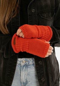 Guanti a maglia senza dita di un vivace arancione con texture a coste. Progettati per una vestibilità aderente e con polsini regolabili. Indossati sopra una giacca nera.