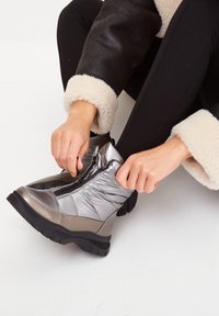 Botte de cheville isolée en argent et gris avec une finition brillante, fermeture éclair sur le côté et semelle noire texturée. Portée avec des leggings noirs et une veste à manche en fourrure.