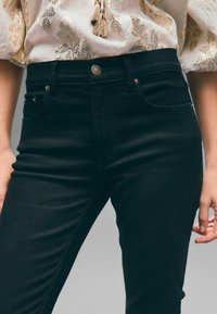 Jeans de mezclilla negra con un diseño ajustado, que cuentan con herrajes plateados, bolsillos frontales y una cinturilla alta. Textura suave y detalles mínimos.