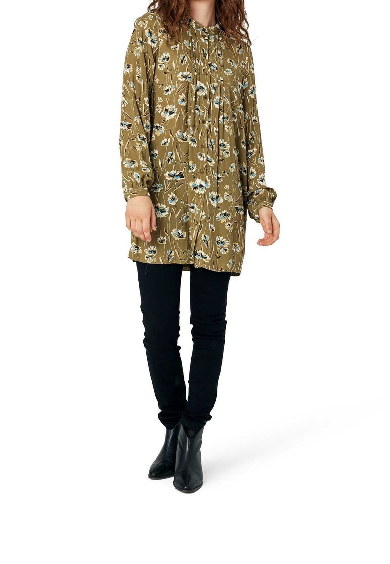 Femme portant une tunique à manches longues à fleurs vert olive, un pantalon skinny noir et des bottines noires, debout devant un fond blanc.