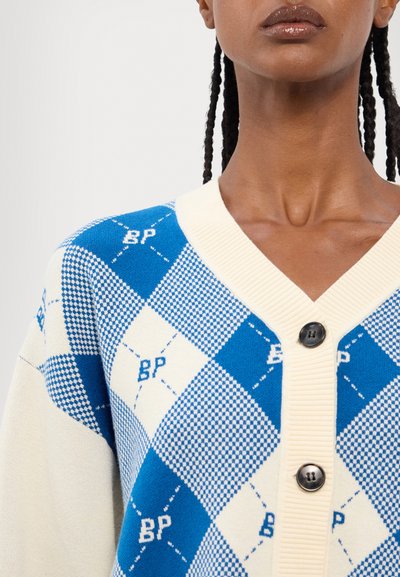 Cardigan bleu et crème avec un motif en losanges, un col côtelé et des boutons noirs. Fabriqué en matériau tricoté doux.