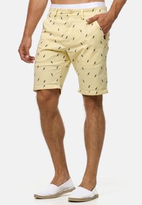 Shorts en coton jaunes avec un motif subtil d'oiseaux bleus, dotés de poches et de revers, associés à des chaussures blanches à enfiler.