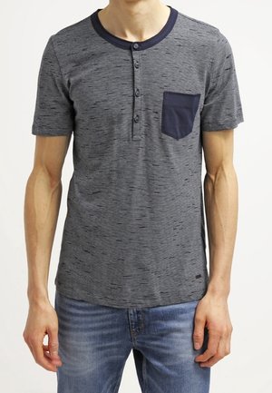 Mand iført en kortærmet grå henley-skjorte med marineblå krave og brystlomme, kombineret med blå jeans.