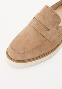 Beige nubukist loafer, millel on õmmeldud disain, kummist väliskreen lainemustriga ja dekoratiivne riba ees.