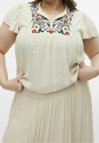 Haut beige clair à manches courtes, avec une broderie florale colorée au niveau du col et une coupe ample. Le tissu est texturé et fluide.