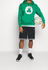 Haut à capuche vert avec logo celtique, shorts noirs avec bord blanc, chaussettes blanches et chaussures de sport noires. Tenant un ballon de basket Nike.