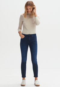 Dunkelblaue Skinny Jeans aus Denim mit hoher Taille und vorderen Taschen, kombiniert mit einem hellbeigen Langarmoberteil und weißen Sneakers.