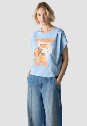 Woman with short blonde hair wearing a light blue T-shirt with orange fruit graphic and text "Freshness to go" and loose blue jeans.