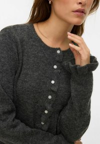 Žena s dlouhými hnědými vlasy, oblečená v tmavě šedém pleteném svetru s malými bílými knoflíky a volánkovými manžetami, dotýkající se krku.