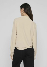 Beige långärmad blus med rynkad krage och manschetter, med en texturerad tygkvalitet. Bärs med svarta byxor som visar en subtil drapering.