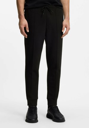 Mężczyzna ubrany w czarne, zwężane spodnie dresowe z troczkiem w pasie oraz czarne trampki, stoi na tle jednolitego białego tła.