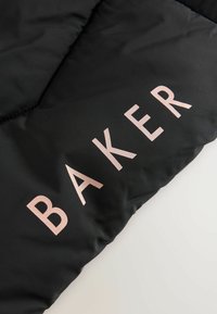 Čierna pufovaná bunda v sekcii s hladkou textúrou, na ktorej je veľký, embosovaný text "BAKER" v ružovom zlate. Výrazný diagonálny vzor šitia.