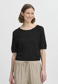 Zwarte gebreide trui met een losse pasvorm, een brede halslijn en korte mouwen met een patroon van kleine gaatjes, gecombineerd met lichtbeige broeken.