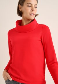 Rotes Langarmoberteil mit hohem Kragen. Glattes, dünnes Material mit taillierter Silhouette. Das Modell hält eine Hand in der Tasche vor einem schlichten Hintergrund.