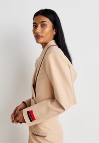 Beige Blazer mit Kapuze, taillierte Passform und Stickerei entlang des Revers und der Taschenränder. Rote und schwarze Akzente am Ärmelbund. Glatte Textur.