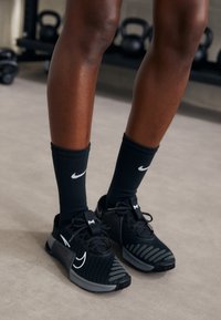Chaussures de sport Nike noires avec une tige texturée, des accents gris et un logo blanc, accompagnées de chaussettes côtelées noires avec un swoosh blanc.