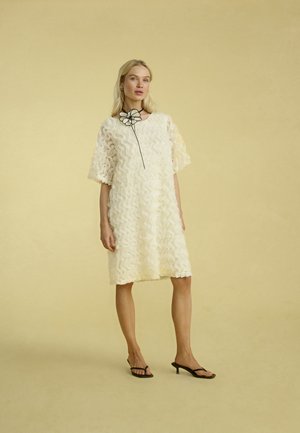 Witte knie-lange jurk met een gewaveerd patroon, losse mouwen en een zwarte bloemenaccent bij de halslijn. Draagt het met zwarte hoge hakken sandalen.