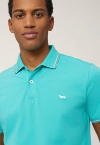 Tirkizna polo majica kratkih rukava, teksturirana tkanina, kontrastne pruge na ovratniku i bijeli logotip jazavičara na lijevoj strani prsnog koša.
