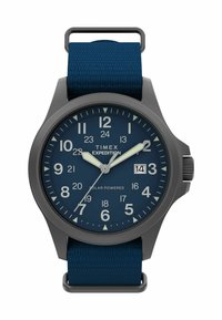 Timex Expedition ur med et mørkegråt urkasse, blå urskive, lysende tal, og et tekstureret blåt stofrem. Har datovindue ved klokken 3.