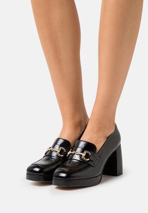 Mocassins à plateforme en cuir noir avec un accent de chaîne dorée et un bout carré, présentant un talon haut chunky et une texture lisse et brillante.
