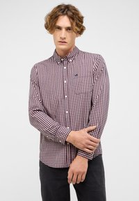 Młody mężczyzna z falującymi włosami nosi koszulę w czerwono-granatowo-białą kratę z guzikami i czarne spodnie, stojąc na tle jednolitego białego tła.