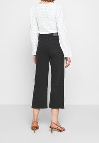 Vaqueros negros de talle alto con corte de pierna ancha, combinados con una camiseta blanca de manga larga con lunares texturizados y sandalias de tacón naranja.