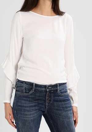Maglietta bianca a maniche lunghe con polsini arricciati, abbinata a jeans blu scuro con chiusura a bottoni e tasche. Tessuto morbido.