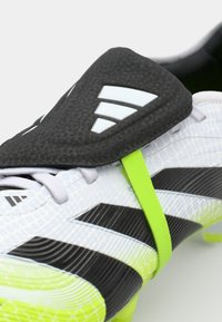 Crampon de football avec une partie supérieure texturée noire et blanche, accentuée de détails en vert fluo, présentant un design épuré et une housse noire protectrice.