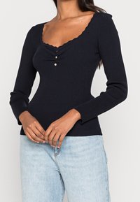 Haut à manches longues côtelé bleu marine avec un décolleté en U festonné, comprenant trois boutons dorés sur la poitrine et une coupe ajustée.