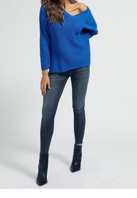 Blauer gestrickter Pullover mit einem schulterfreien Design, langen Ärmeln und einem strukturierten Muster, kombiniert mit enganliegenden dunkelgrauen Jeans und schwarzen Knöchelstiefeln.