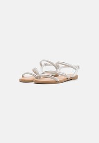 Flat sandals with multiple silver straps and a beige sole, featuring a simple buckle closure and a smooth, textured surface.