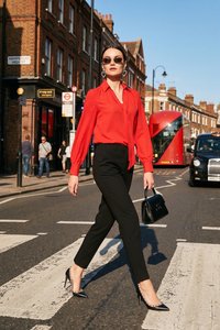 Femme en blouse rouge et pantalon noir marche avec assurance dans une rue de Londres, tenant un sac à main noir, avec un bus à impériale et un taxi derrière elle.