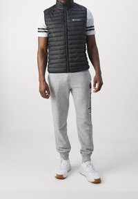 Černá péřová vesta s vysokým límcem, šedé tepláky s modrým logem a bílé tenisky. Na tričku pod vestou jsou vodorovné pruhy.