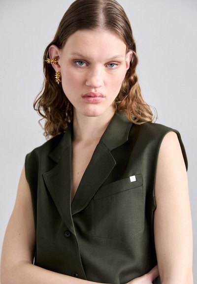 Aliejinės žalios spalvos be rankovių švarkas su iškirpimu, vienu krūtiniu kišeniu ir metaliniu logotipo akcentu. Glotni audinio tekstūra, mygtukų detalės.